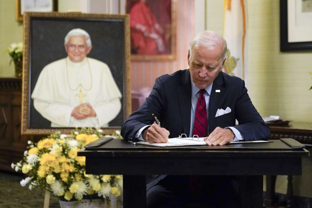 Biden sits at a desk and signs a book with a portrait of Pope Benedict XVI in the background