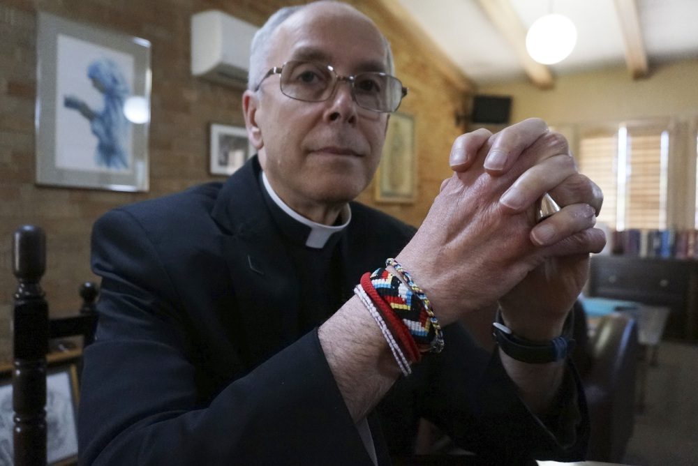 A white bishop wearing glasses and brightly colored friendship bracelets folds his hands