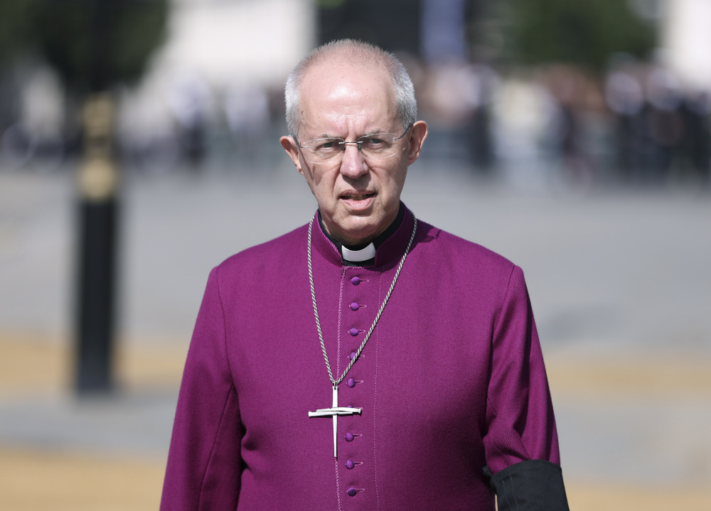 An older white man with glasses wears a magenta shirt, a clergy collar, and a cross necklace