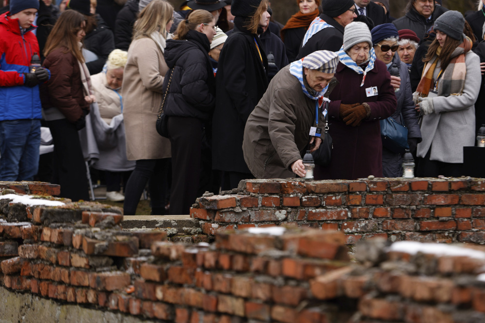 Two older wearing light blue and white striped handkerchiefs around their necks lay candles on a brick wall with a crowd behind them