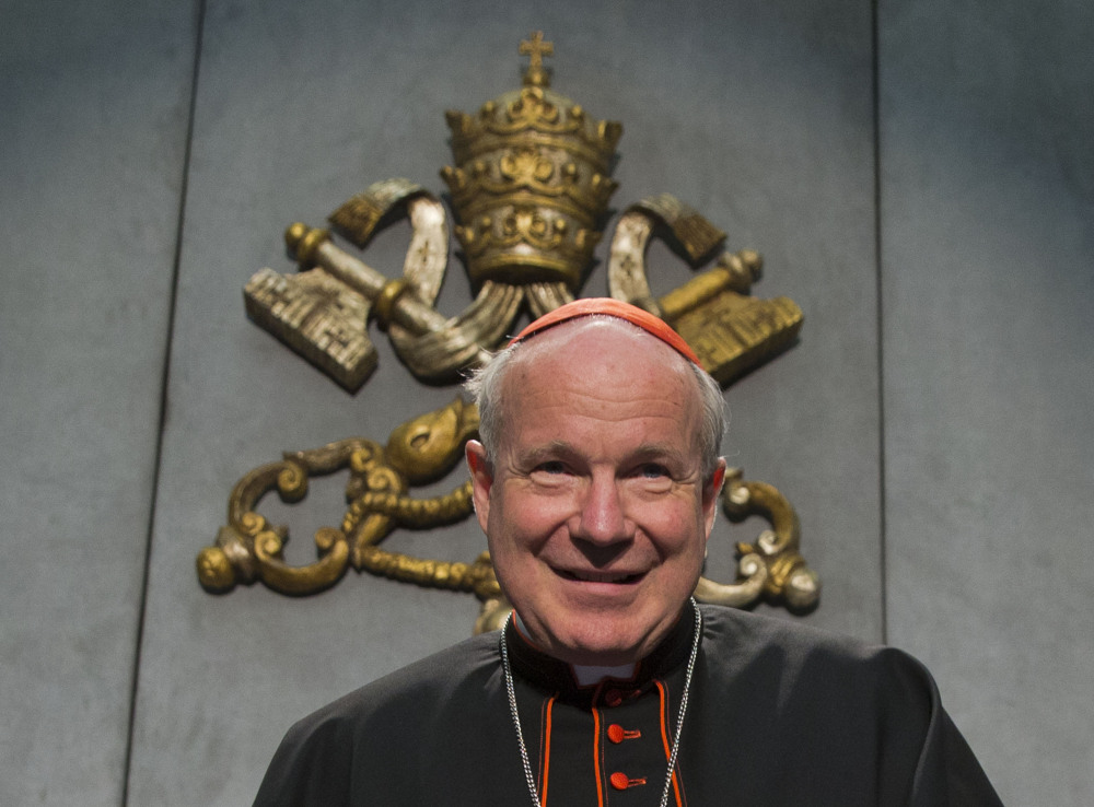 An older white man wears a red zucchetto and clergy collar while sitting in front of the Vatican crest