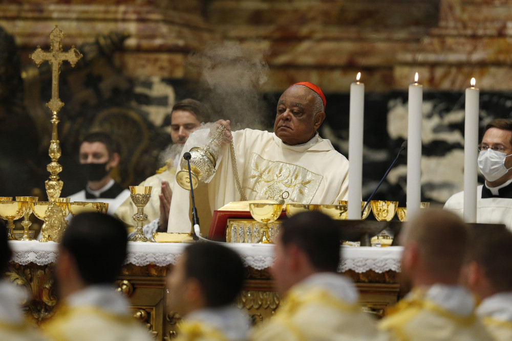 A Black man wearing a red zucchetto disperses incense behind a large altar