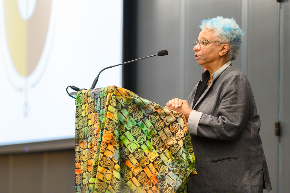 An older Black woman with blue streaksin her hair speaks at a podium covered in a brightly colored cloth