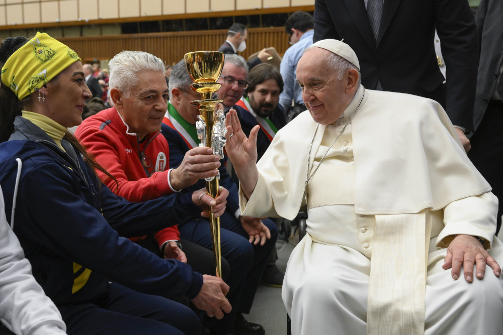 Pope Francis, sitting, blesses a gold torch held by a man and a woman