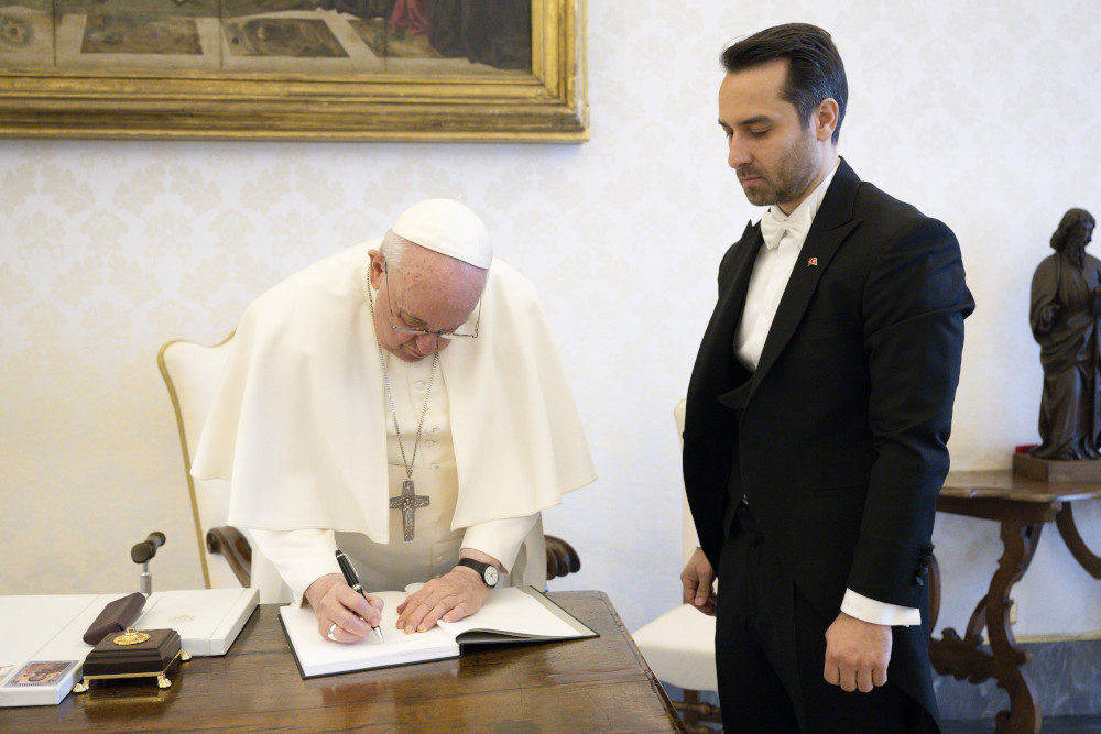 Pope Francis writes in a notebook on a desk. Next to him stands a man in a tuxedo