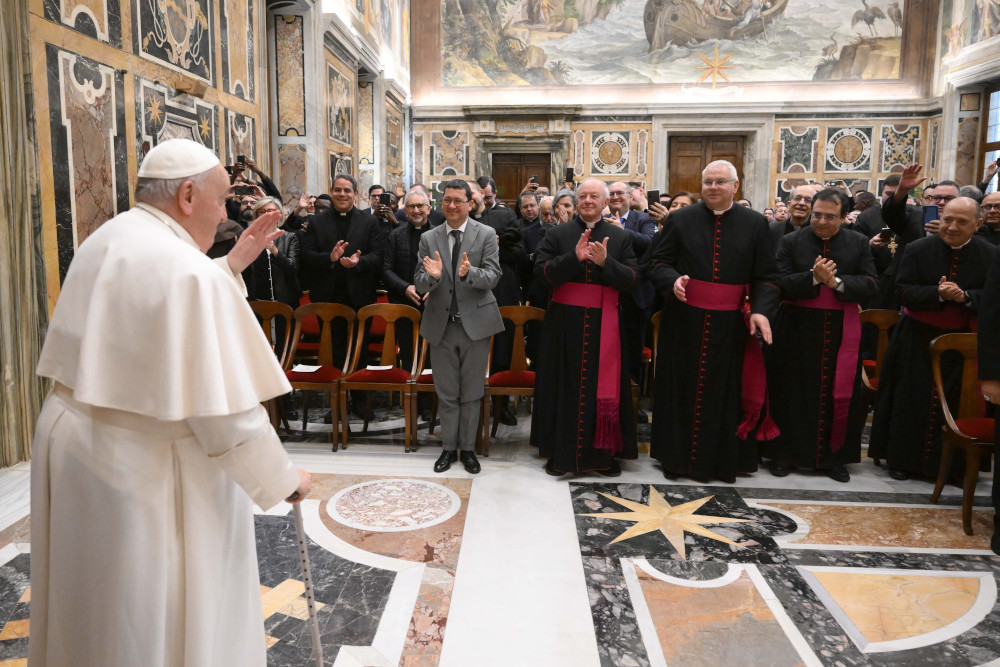 Pope Francis stands in front of and holds his hand in a blessing gesture towards a large audience of men, mostly clerics