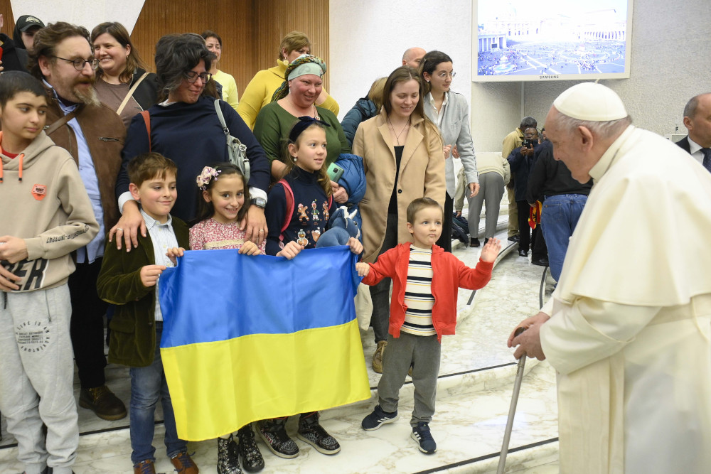 Pope Francis, using a cane, speaks with a group of children holding a Ukrainian flag and their adults