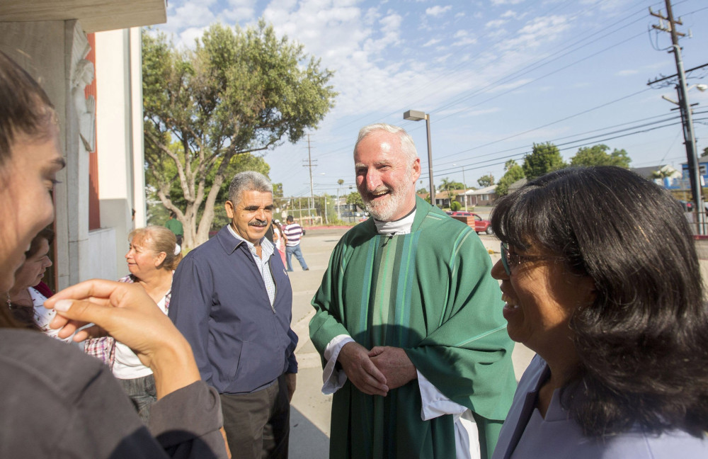 A white man with white hair wears green vestments and talks to a group of people outside