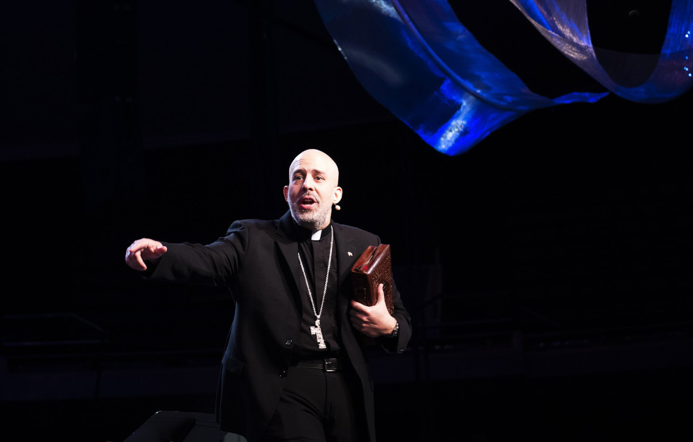 A bald man in a black cassock and a pectoral cross gestures energetically on a dark stage