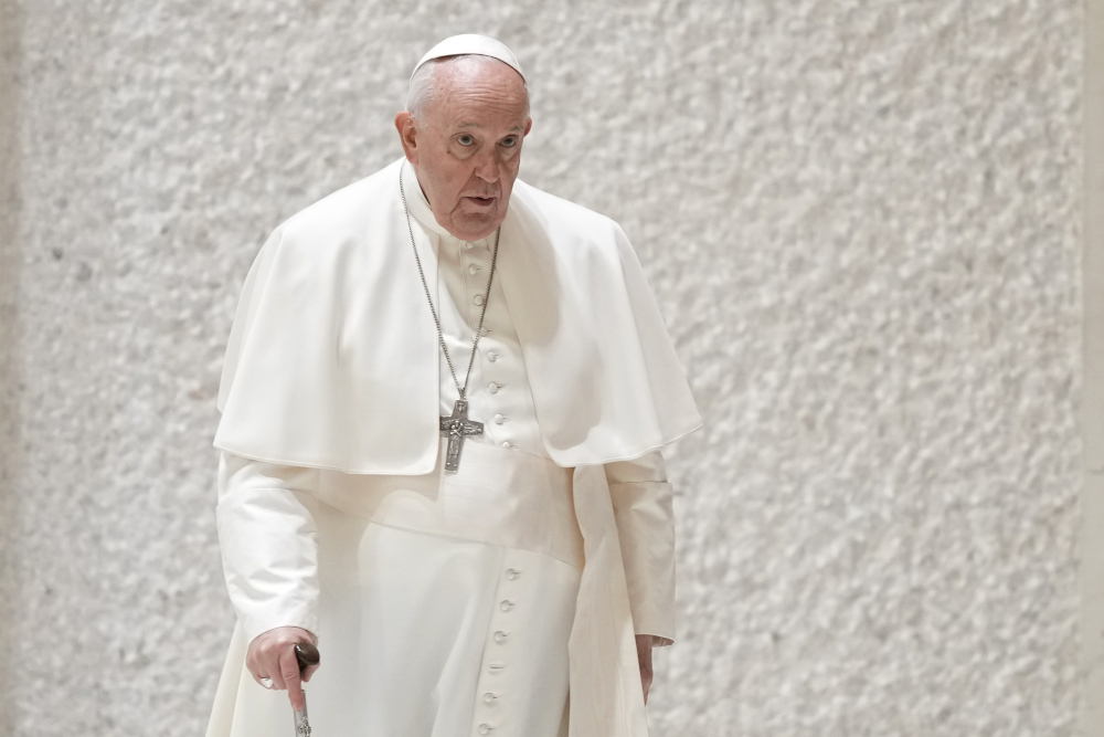 Pope Francis walks with a cane by a white wall