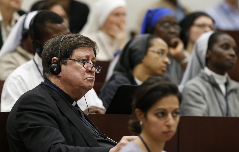 A man in a black cassock wears headphones and sits among women religious and other people
