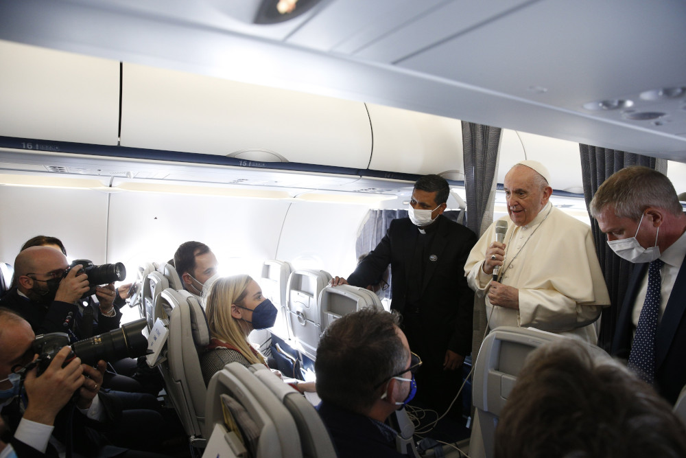 Pope Francis stands in the middle of a plane and speaks with masked journalists who surround him