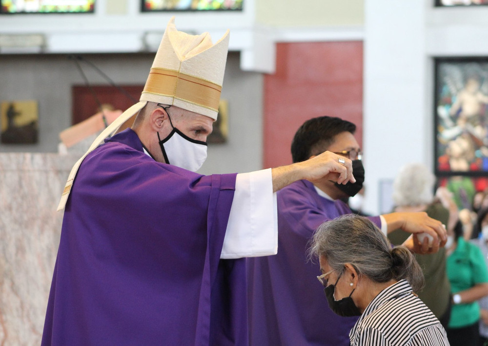 A man wearing a mitre, a mask, and purple robes sprinkles ashes over the head of a woman wearing a mask