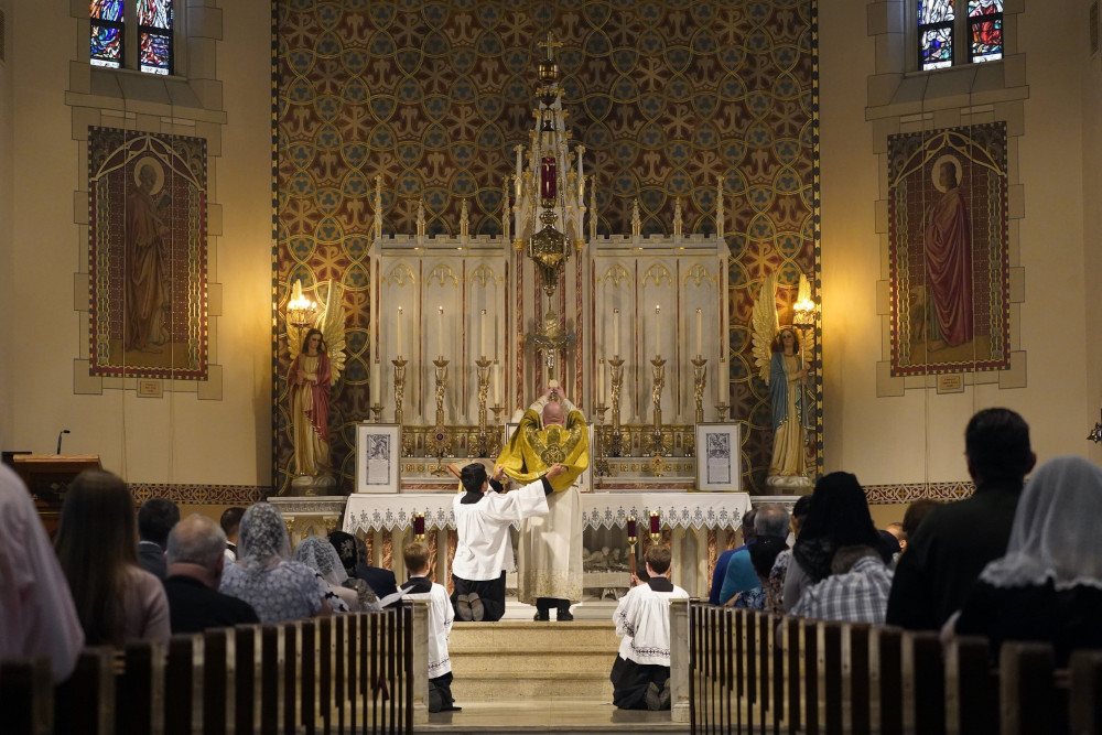 A priest with his back to the pews full of people, where some women wear lace veils, lifts a Communion host high