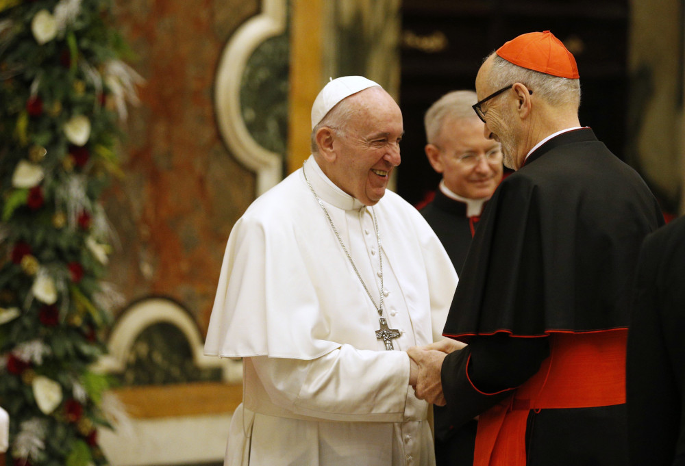 Pope Francis shakes hands with an older white cardinal. Another man is in the background