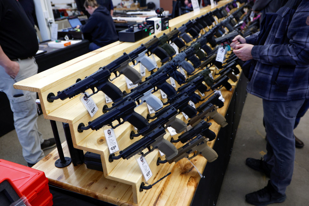 Rows and rows of guns are displayed on shelves. The body of a potential gun buyer is visible.