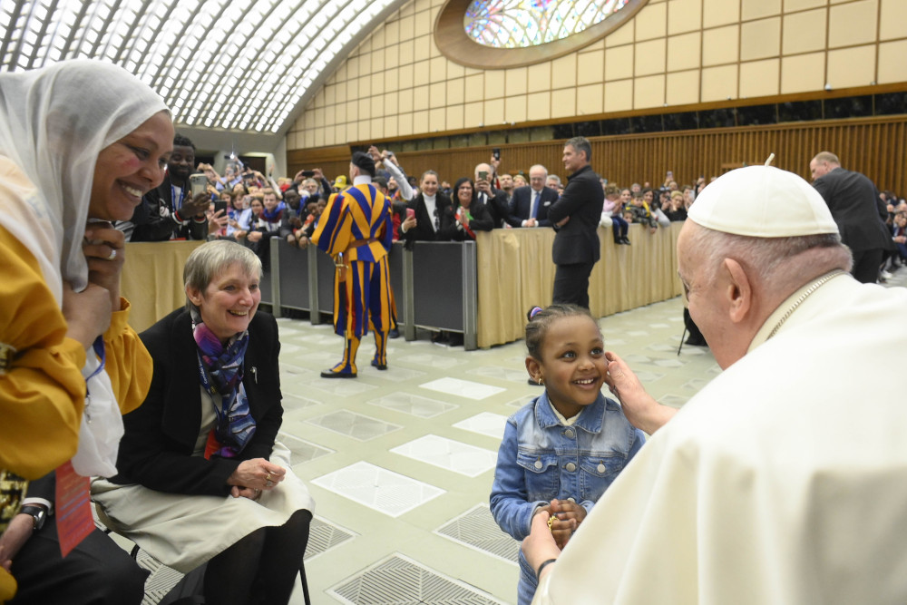 Pope Francis touches the cheek of a smiling Black girl. A short-haired white woman and Brown woman with her hair covered smile as they watch.