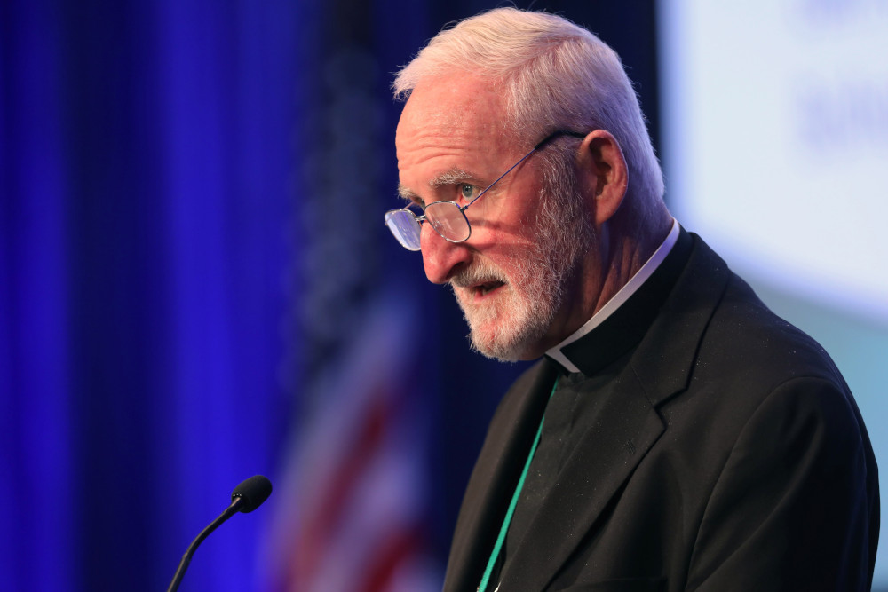 A white man with white hair, a beard, glasses, and a black cassock speaks into a microphone