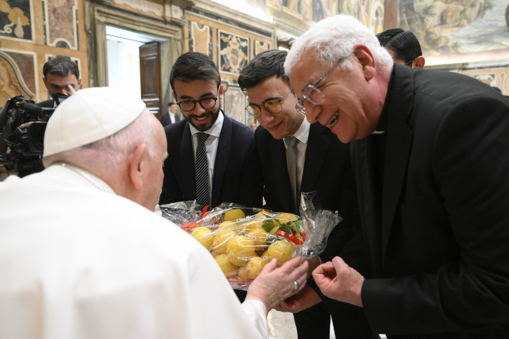 A group of men in suits and cassocks gather around Pope Francis and hand him a seran-wrapped container of lemon-looking fruit