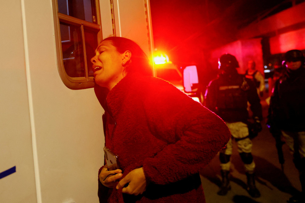 A woman leans on an ambulance in severe distress, clutching a cellphone, in red emergency light