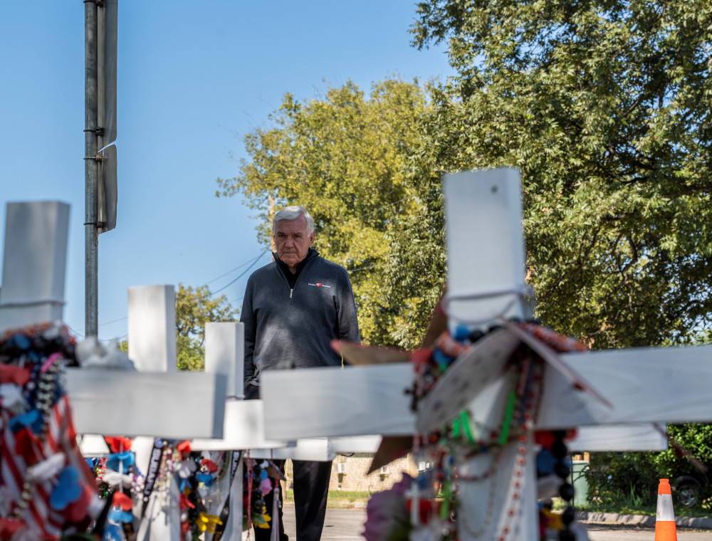 An older man in a quarter zip sweater stands before white crosses outside
