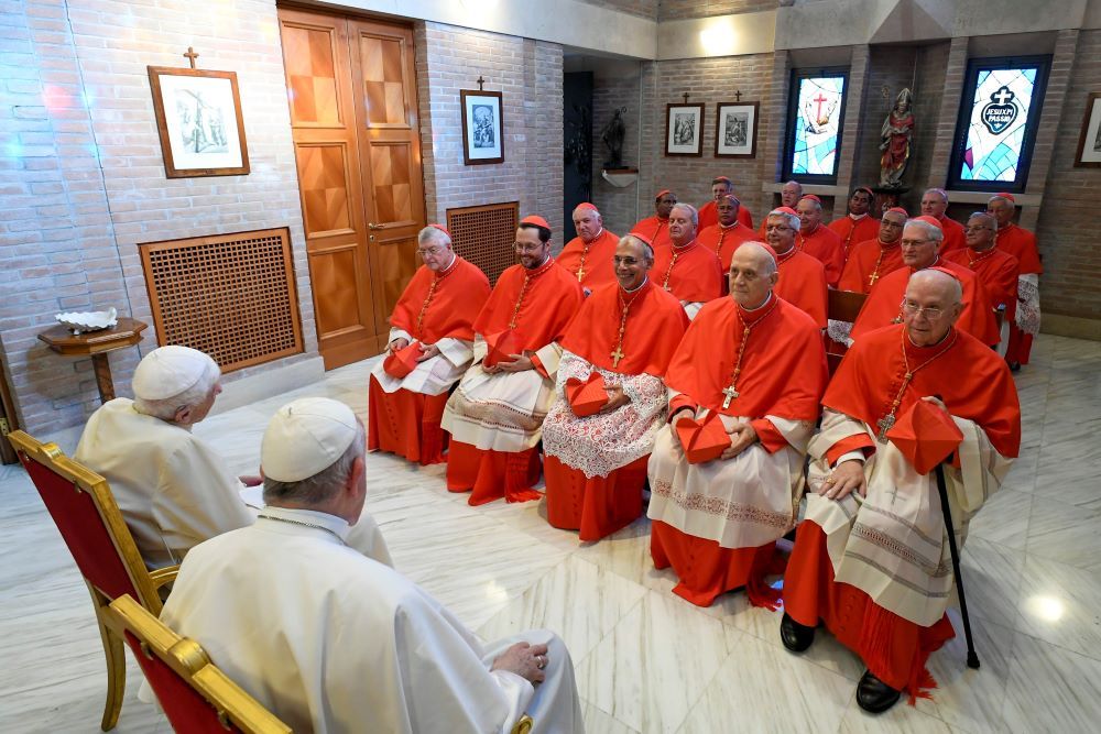 New cardinals, dressed in red, sit and talk with Pope Francis and Pope Emeritus Benedict XVI.