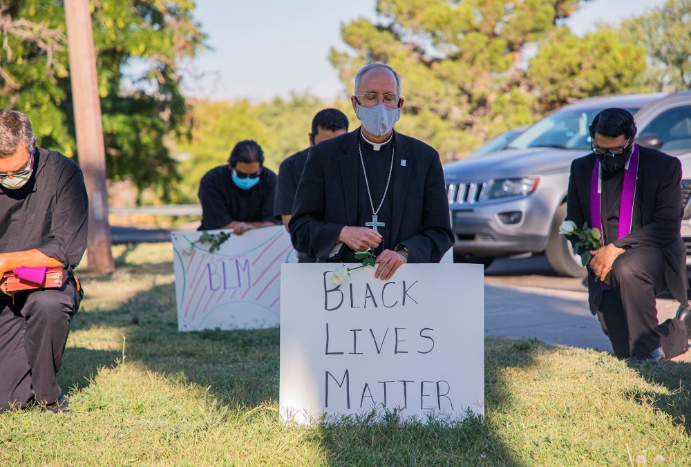 Bishop Mark Seitz kneels, wearing a mask and holding a sign that says "Black Lives Matter."