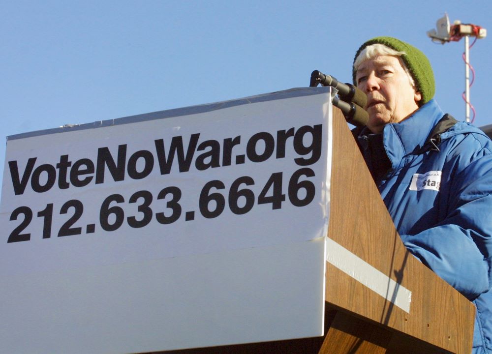 Woman speaks at lectern with sign that reads, "VoteNoWar.org."