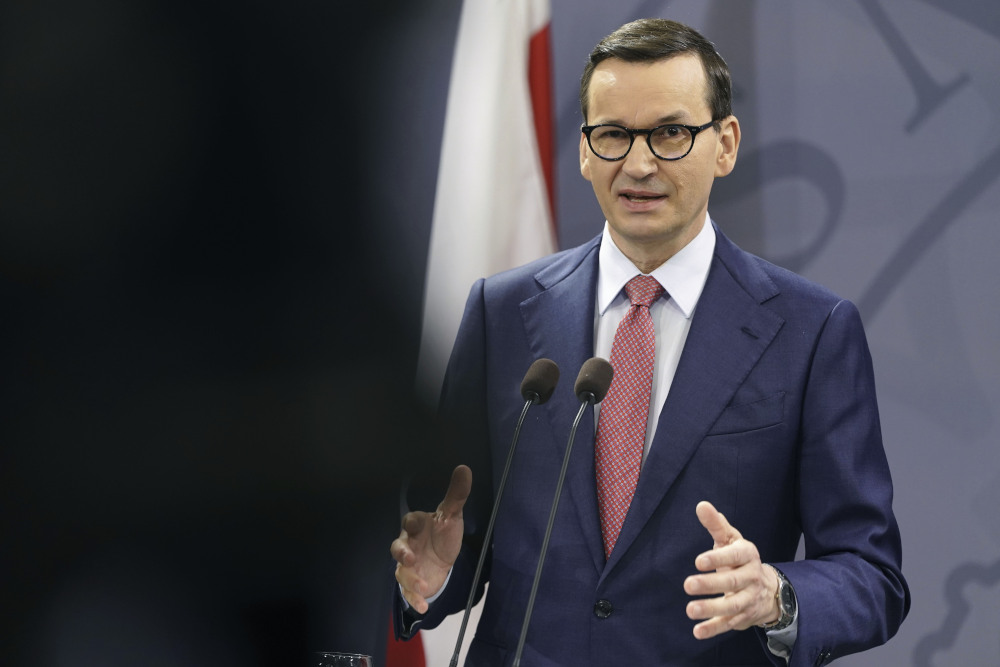 A skinny white man with glasses and a suit makes a gesture with his hands and speaks into a microphone