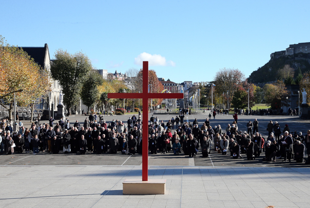 A large group of people dressed in black kneel in front of a large red cross outside
