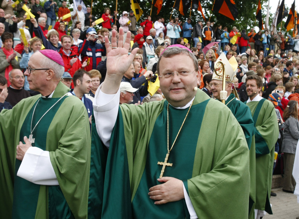 A white man wears glasses, a green robe, a violet zucchetto, and a pectoral cross and waves at a parade audience