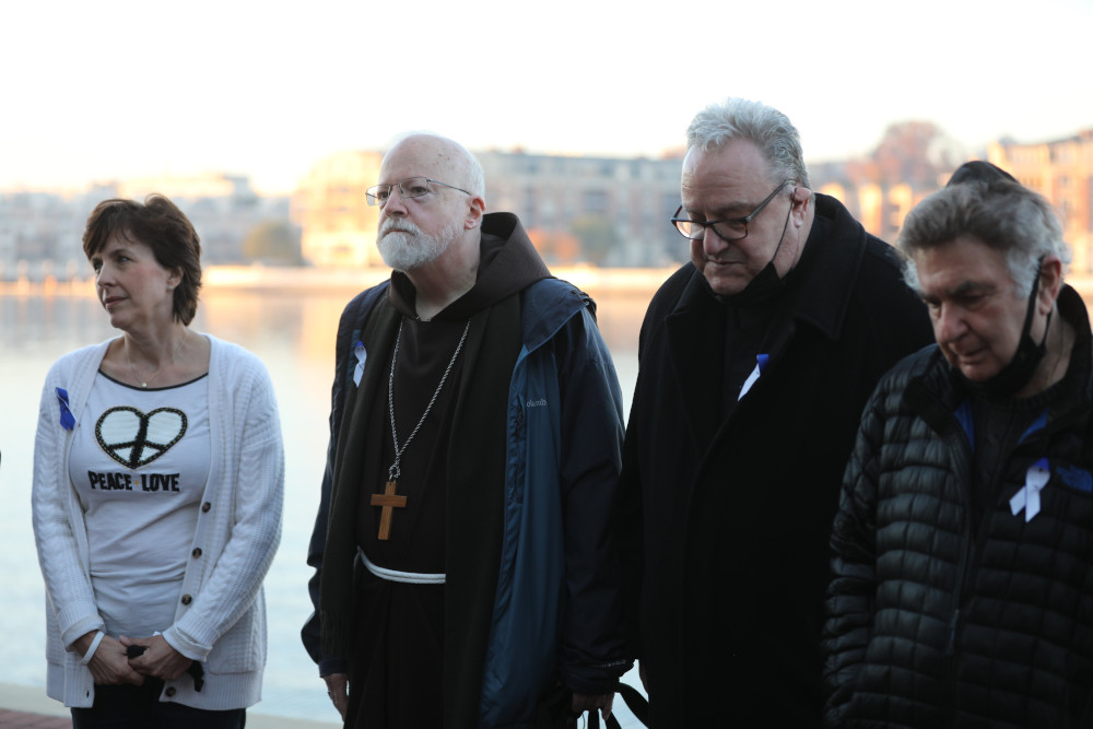 An older white man wears a brown robe and a wooden pectoral cross as he stands between two men and a woman