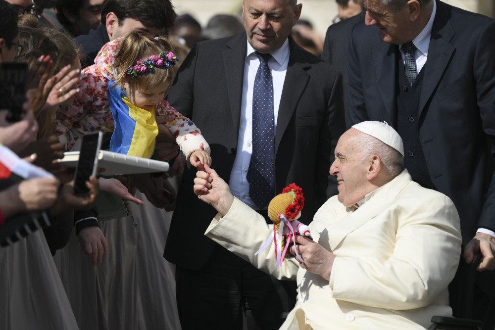 Pope Francis holds a little girl's hand. The little girl is wearing a Ukrainian flag. Pope Francis holds a doll wearing a flower crown.