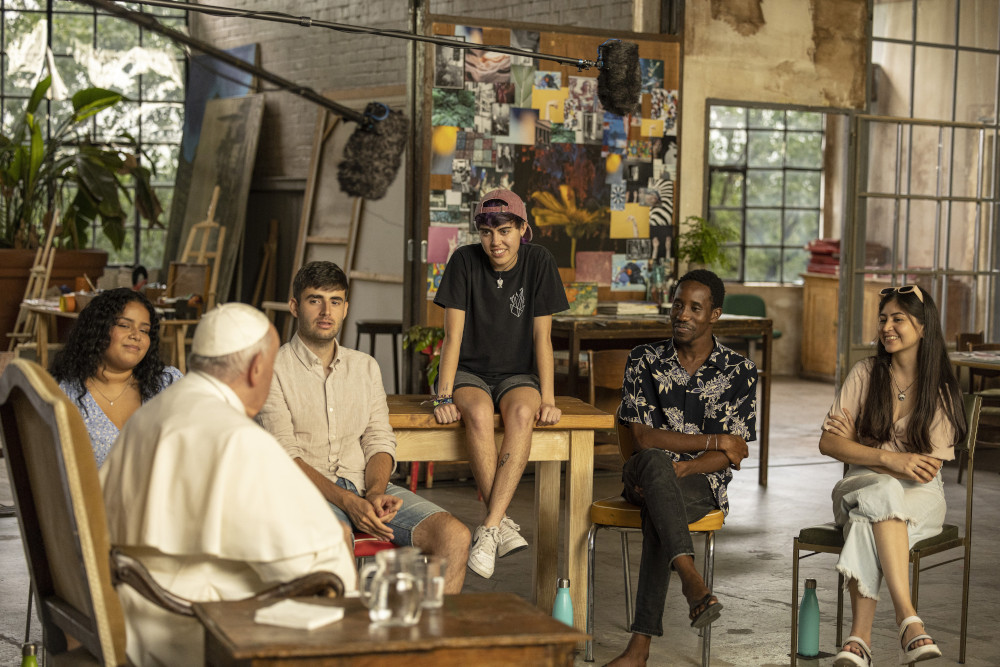 Pope Francis sits with a group of young adults in a circle in what appears to be an artists' loft. A boom mic is visible, and one young adult perches on a table.