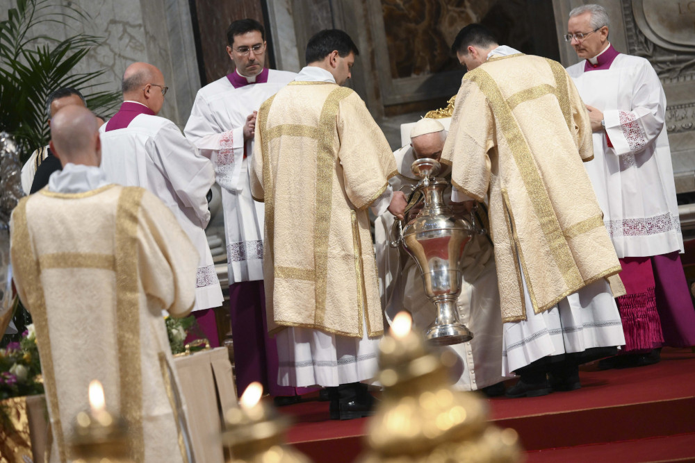 Pope Francis bends over a silver urn, while surrounded by men in white and golden vestments
