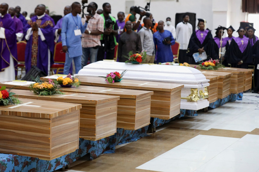 A row of caskets have flowers placed on them. They are surrounded by people in various church garments and other people with news cameras.