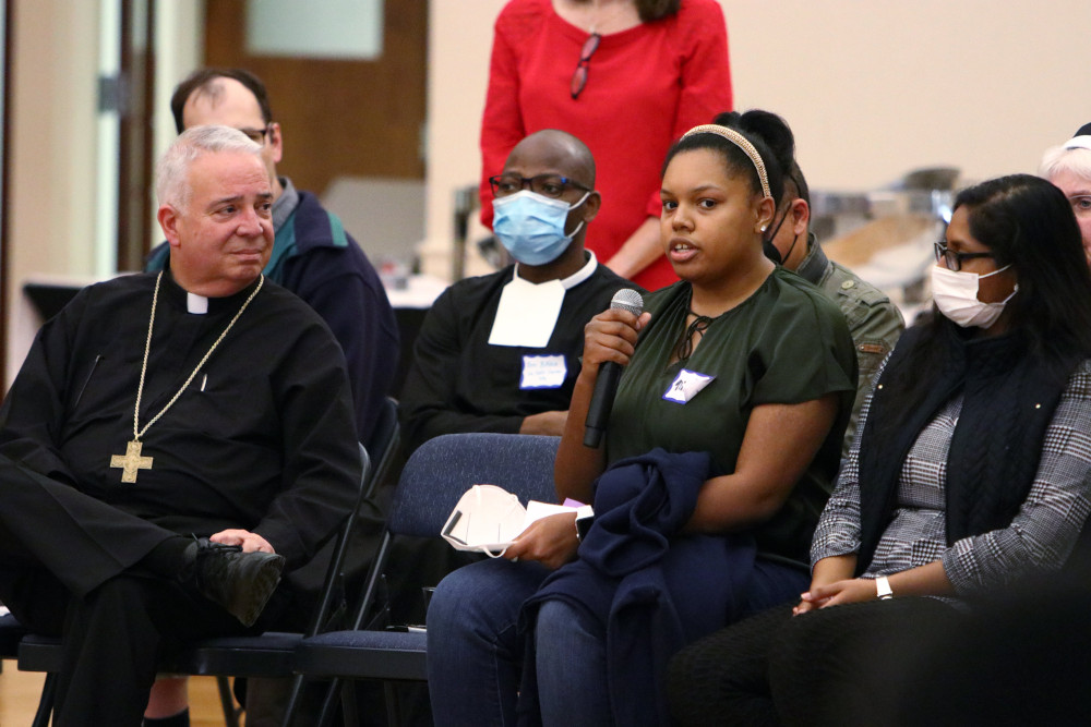 A man wearing a black cassock and a pectoral cross looks on as a young Black woman speaks into a microphone, while surrounded by other people in chairs, some masked