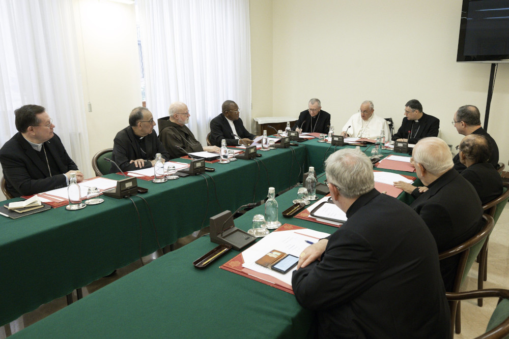 A group of men in black cassocks sits at long tables in a U shape with Pope Francis, without his zucchetto, at the head