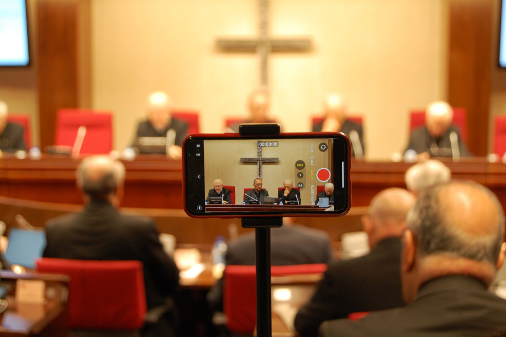 A phone captures video of a press conference led by several clerics in front of a crucifix