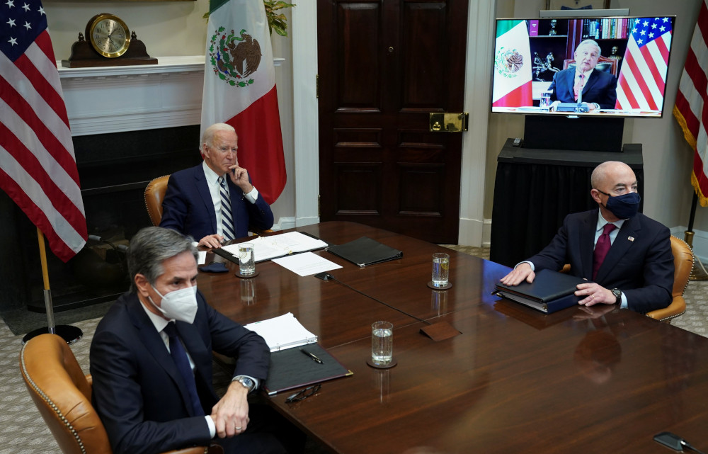 Two men in suits and masks sit on either side of Joe Biden, also in a suit, at a long table. A man with a Mexican and US flag behind him is on a video screen.