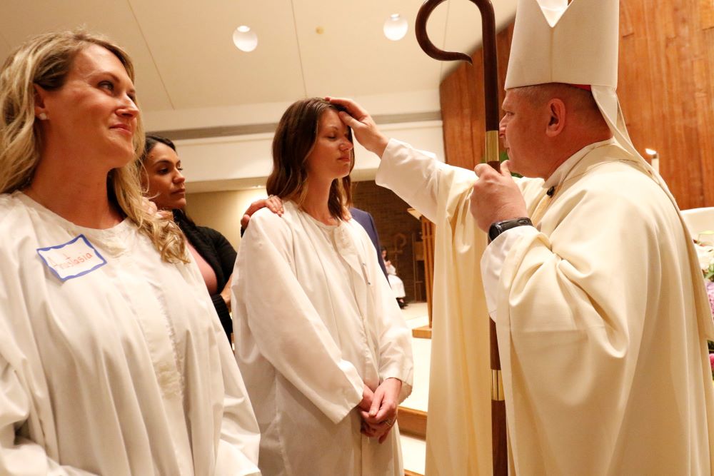 Bishop makes sign of cross on forehead of young woman, while another woman looks on.