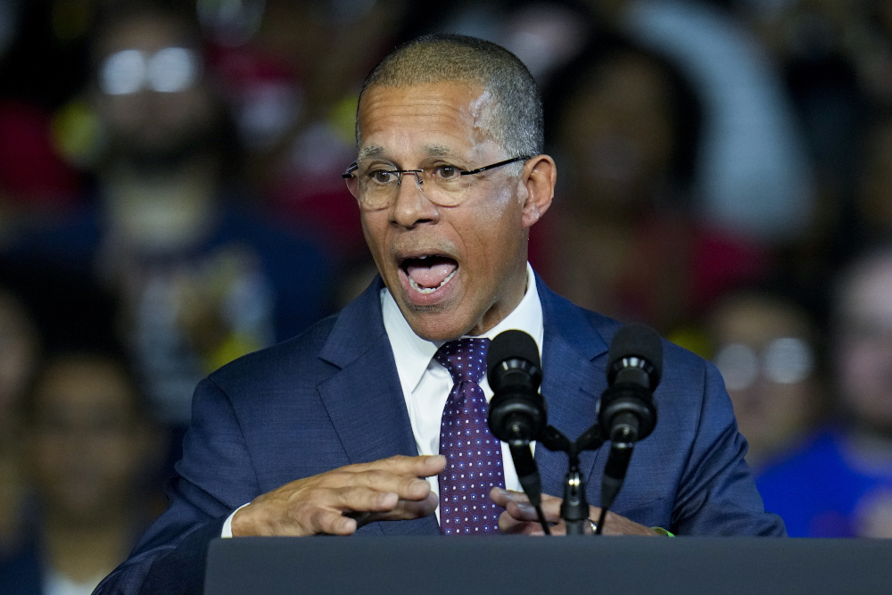 A light-skinned Black man wearing glasses and a suit speaks into a microphone at a political rally