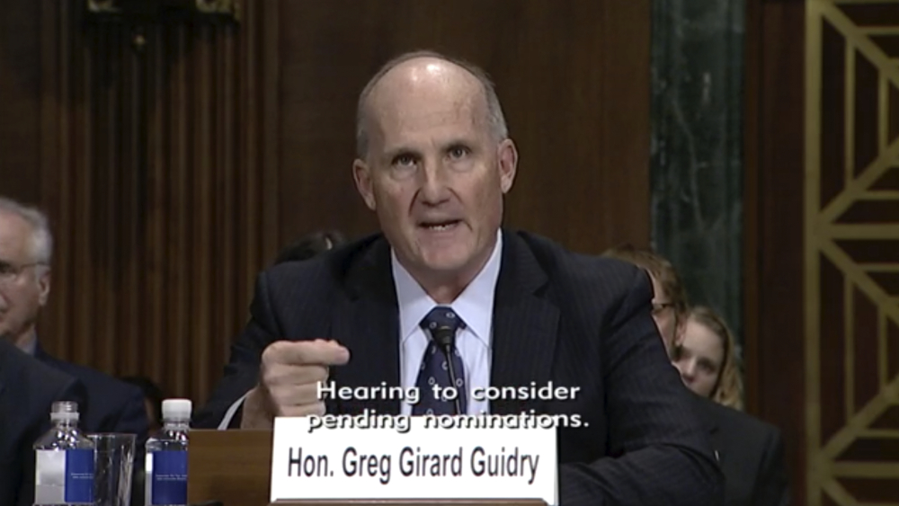 A bald white man in a suit sits behind a paper placard that reads "Hon. Greg Girard Guidry"