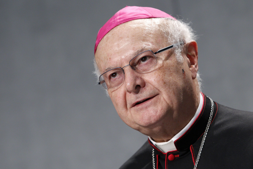 An older white man wearing glasses wears an amaranth zuchetto along with a black cassock