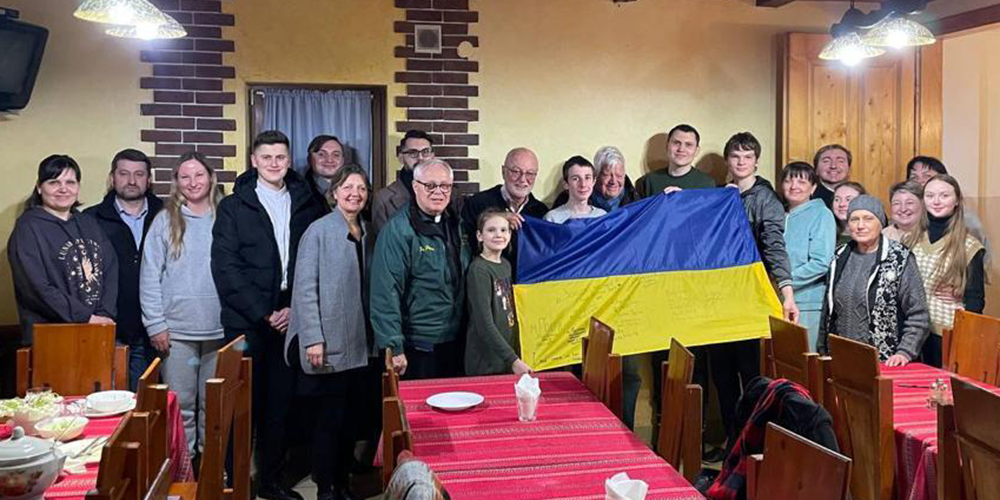 Refugees at the Catholic retreat house in the Carpathian Mountains in Ukraine are pictured with visitors from the U.S., including NCR contributor Fr. Peter Daly, front row, sixth from the left, NCR board member David Bonior, holding the flag, and Jim McDermott, to his left. Refugees signed and presented a Ukrainian flag to the delegation. (Courtesy of David Bonior)