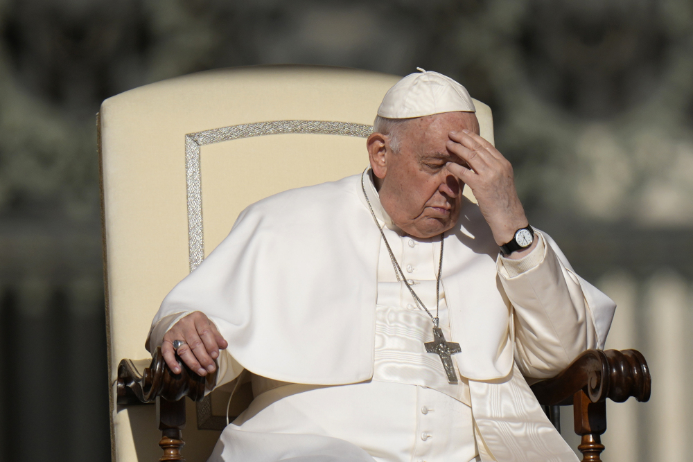 Pope Francis, sitting in an ornate cream chair, touches his forehead as if making the sign of the cross