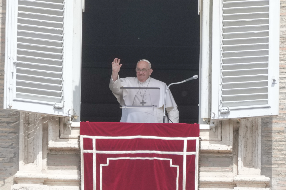 Pope Francis waves from the window of his papal apartment while standing behind a clear lecturn