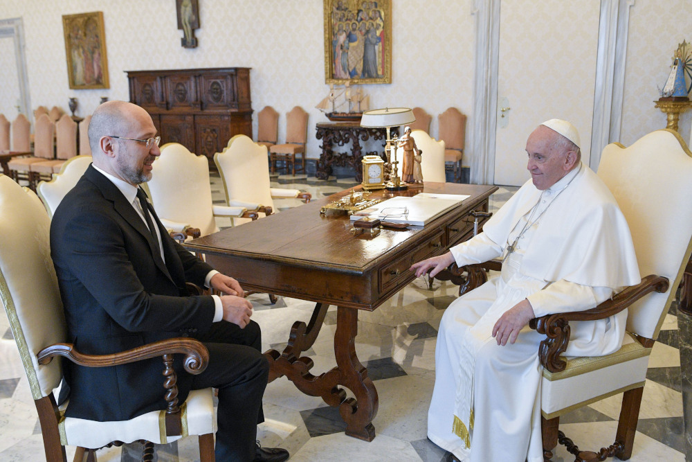 A white, bald man in a suit sits across from Pope Francis at the corner of a table