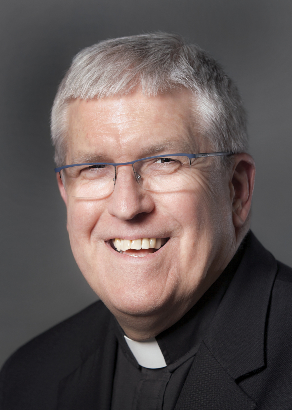 A white man with white hair and glasses wears a cassock and smiles into the camera