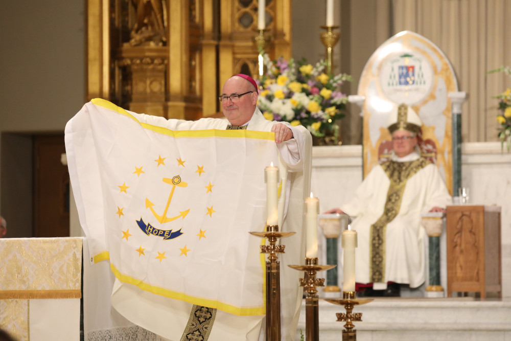 A white man with an amaranth zucchetto holds a Rhode Island state flag in front of a cathedral. A white man with a mitre is visible in the background.